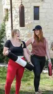 Two ladies modeling lululemon outside of Hill Country Herb Garden Spa. The woman on the left is holding a yoga mat, wearing red yoga pants and a black, short-sleeve crop top. Te woman on the right is wearing black yoga pants, a maroon tank top and holding a red lululemon water bottle.